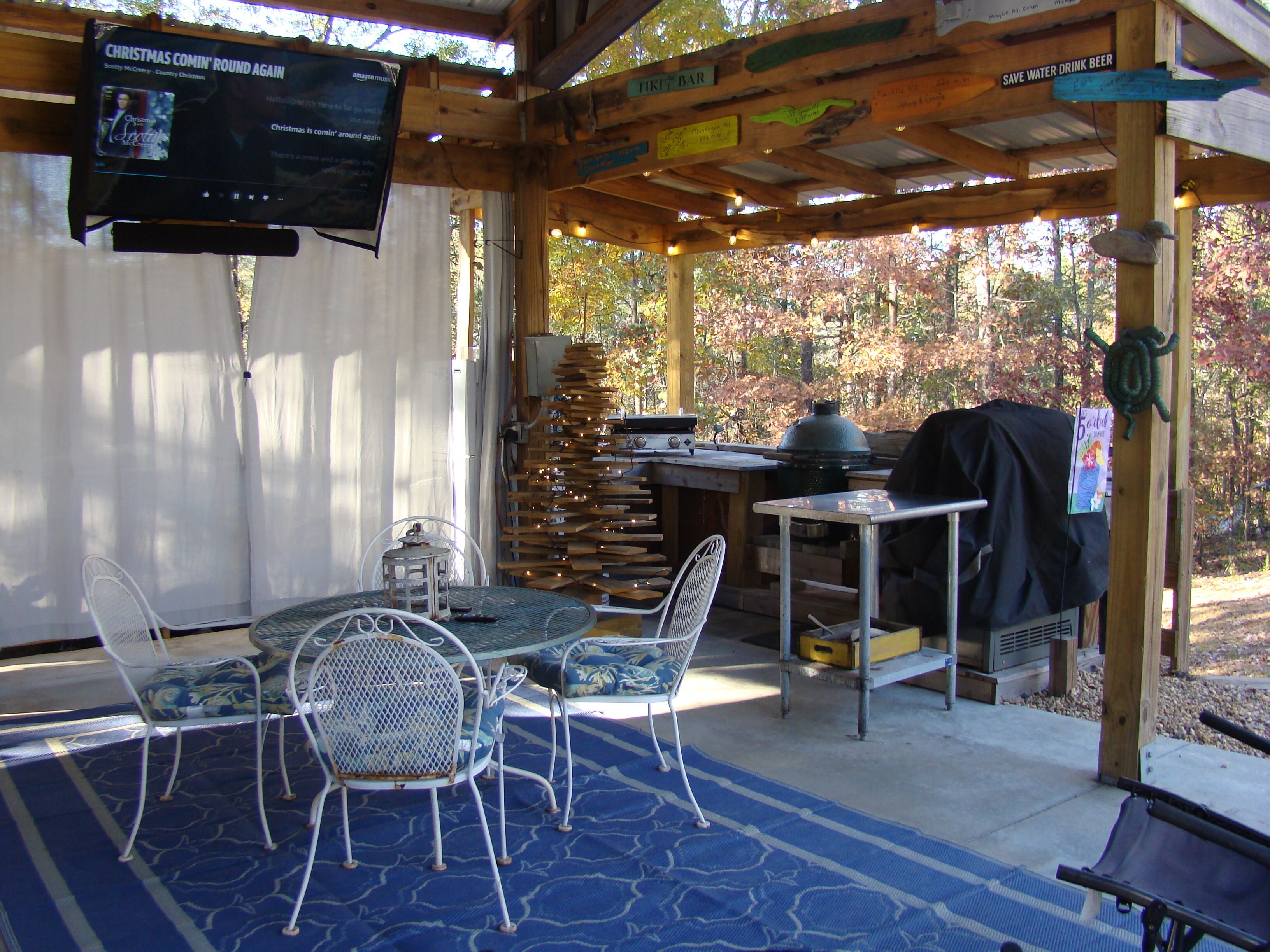 Portico esterno coperto con schermo televisivo, mobili in legno, barbecue e un cartello che invita al risparmio idrico.
