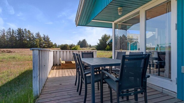 A wooden deck with a table and chairs, overlooking a grassy field and trees. - 5 Star Holiday Home in Blokhus (Blokhus)