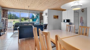 A cozy room with a wooden ceiling, a dining table with chairs, a sofa, and a view of a blue house through large windows. - 5 Star Holiday Home in Blokhus (Blokhus)
