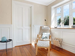 A room with a wooden floor, white walls, a white door, a window with a view, a chair with a cushion, and a small table with a blue object. - 4 Star Holiday Home in Lokken (Lokken)