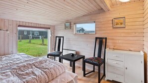 A cozy cabin room with a bed, a small table, two chairs, a cabinet, and a window overlooking a grassy yard. - 5 Star Holiday Home in Lokken (Lokken)