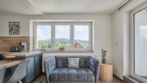 Ett modernt kök med en grå soffa, ett matbord och stolar. Det finns ett fönster med utsikt över träd och kullar.
