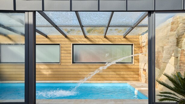 En modern inomhuspool med väggar av genomskinligt glas, träpanel och en vägg av sten.