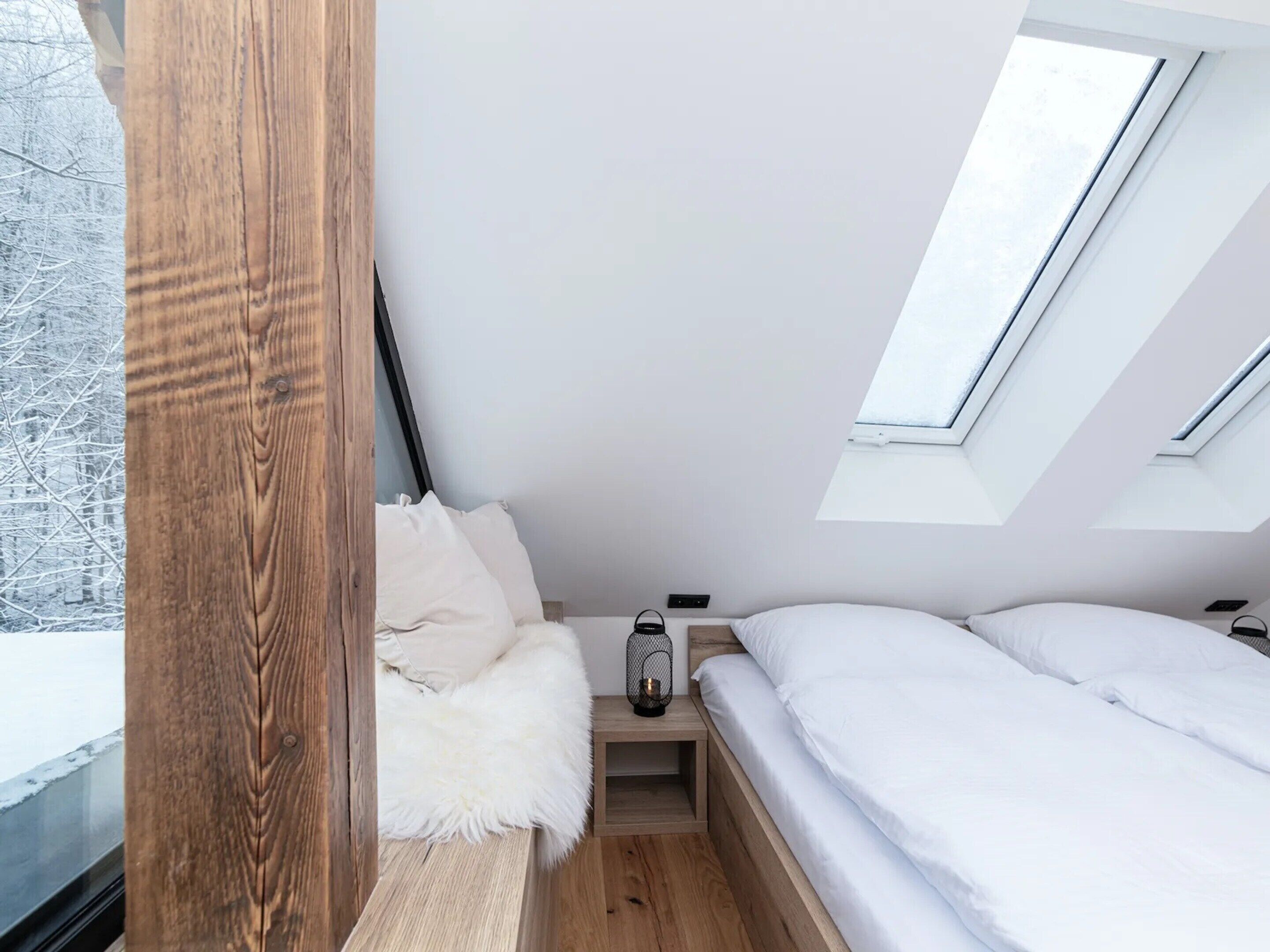 A compact, modern bedroom with a sloped ceiling, skylights, a wooden bench, and a small bedside table.
