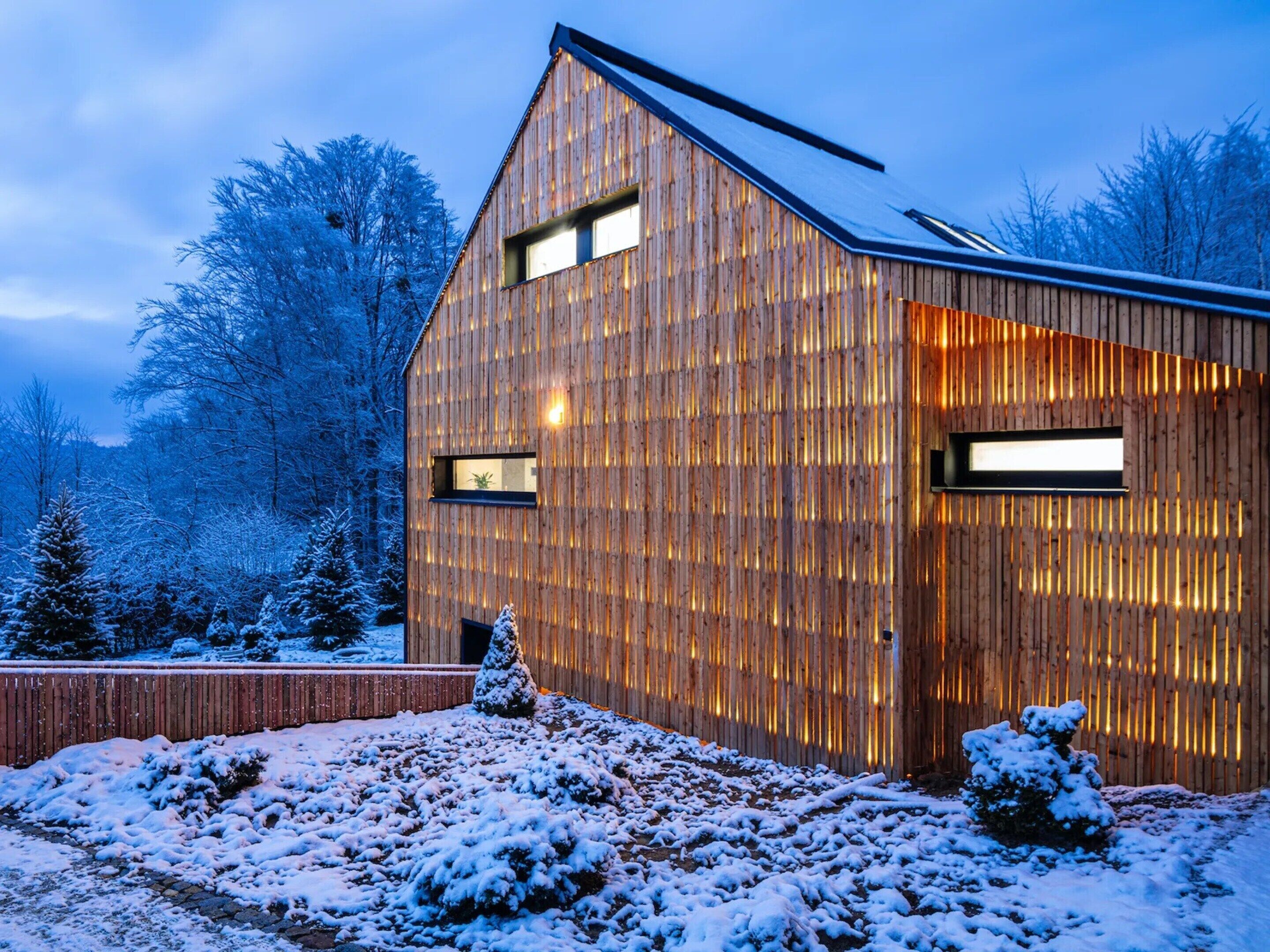 Ein modernes Holzgebäude mit großen Fenstern und einem geneigten Dach, umgeben von schneebedeckten Bäumen.