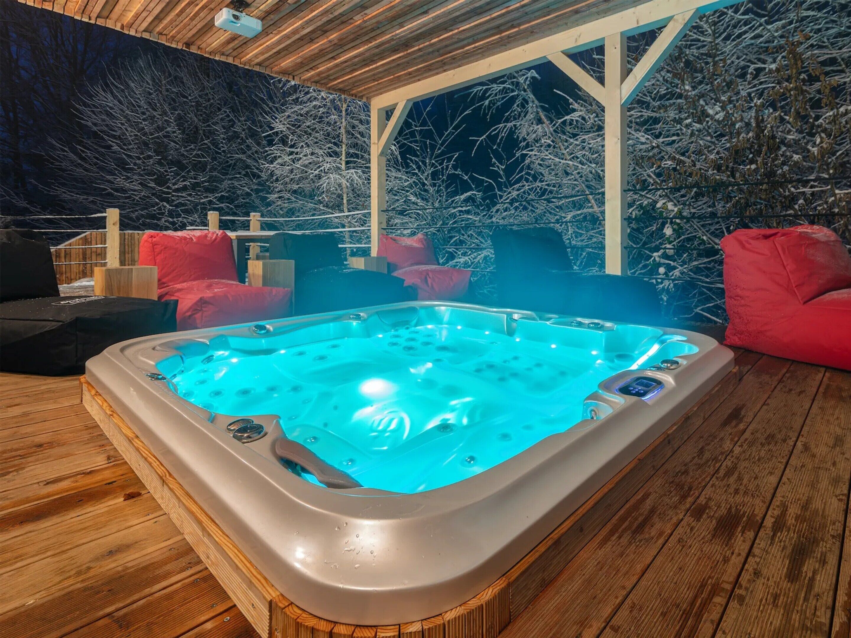 A hot tub with a wooden deck and red cushions, surrounded by snow-covered trees.