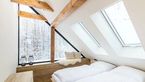 A bedroom with a sloped ceiling, wooden beams, a bed, a bench, and a view of a snowy forest through large windows. - Villa in Celadna With Outdoor Cinema & Whirlpool (Celadna)