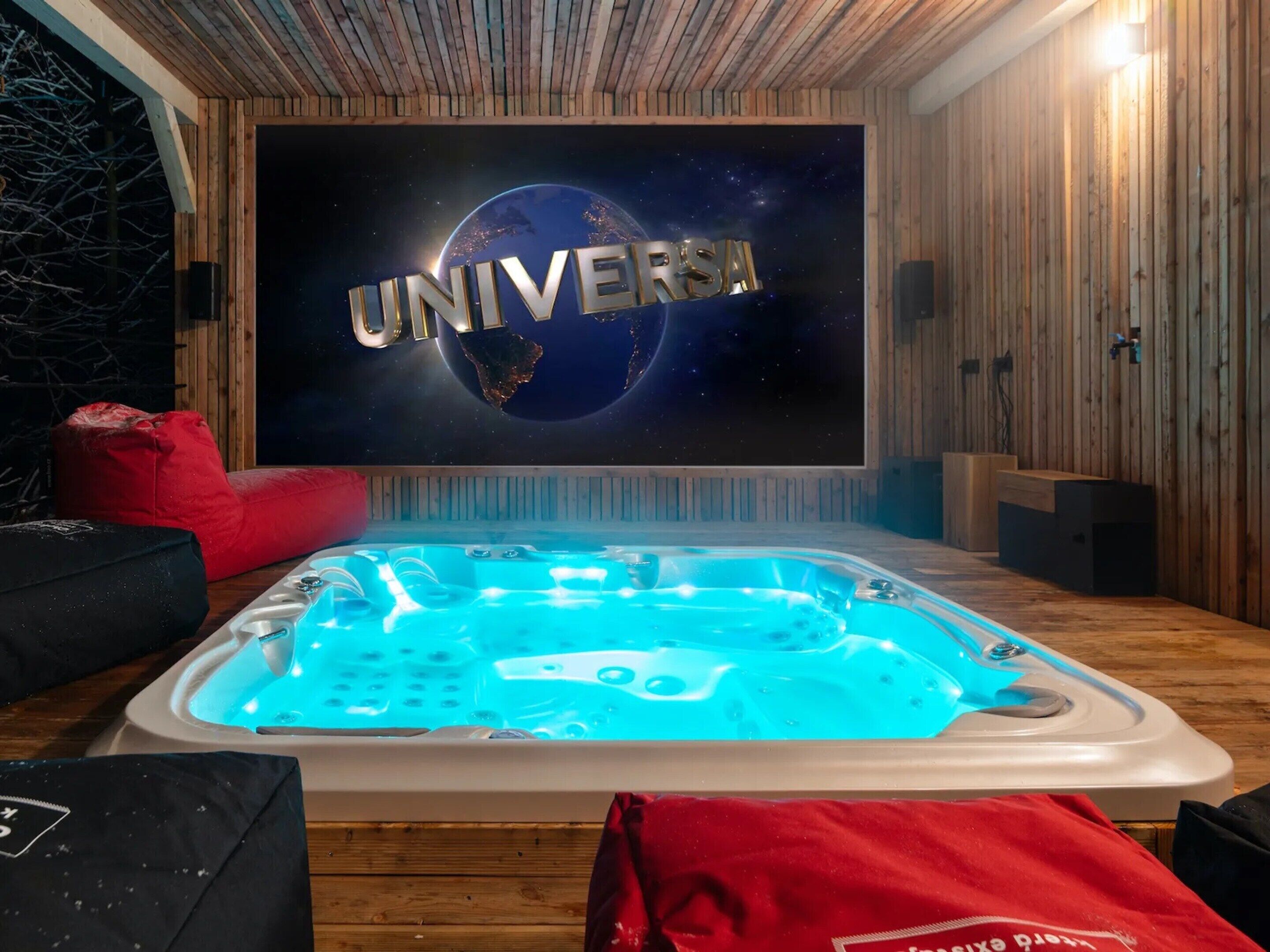 Une pièce avec un décor intérieur en bois, un grand écran affichant le mot « UNIVERSAL », un jacuzzi éclairé de lumières bleues et un fauteuil en forme de sac de haricots rouge.
