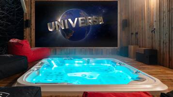 Une pièce avec un décor intérieur en bois, un grand écran affichant le mot « UNIVERSAL », un jacuzzi éclairé de lumières bleues et un fauteuil en forme de sac de haricots rouge.