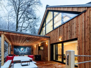 A wooden cabin with a large glass door, a covered outdoor seating area, and a poster on the wall. - Villa in Celadna With Outdoor Cinema & Whirlpool (Celadna)