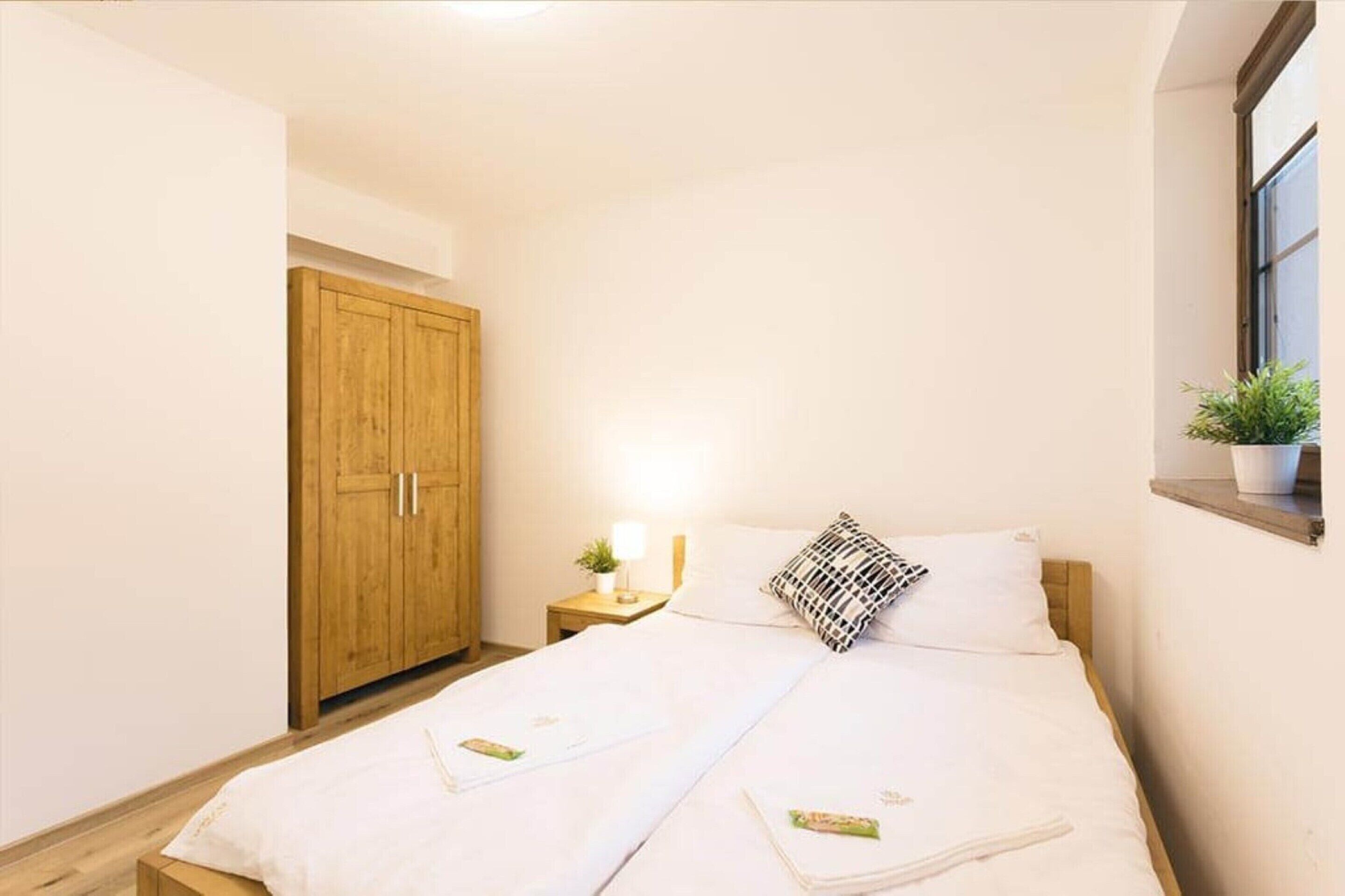 Un dormitorio con dos camas, un armario de madera, una mesita de noche con lámpara y una ventana con una planta.