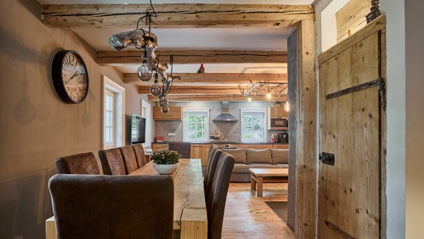 A dining area with a wooden table and chairs, a clock on the wall, and a view into the kitchen area with wooden beams and a large wooden door. - Chalet in Horni Marsov Near Ski Slopes (Horni Marsov)