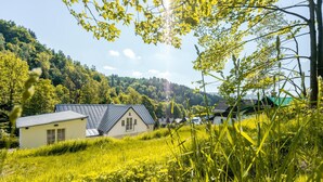 Ein Haus mit geneigtem Dach und einer kleinen Veranda, umgeben von üppiger Vegetation und einem bewaldeten Hügel im Hintergrund.