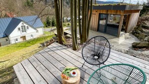 Eine Holzterrasse mit zwei Stühlen, einem kleinen Tisch mit einem Strauß Tulpen und einer Teetasse, mit Blick auf ein Haus und Bäume im Hintergrund.