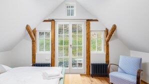 Ein Schlafzimmer mit schrägem Dach, Holzbalken, einem Bett, einem Sessel und großen Fenstern mit Blick auf Grünflächen.