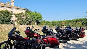 Motorräder sind auf einer Schotterfläche abgestellt, im Hintergrund sind ein Gebäude und Grünflächen zu sehen.