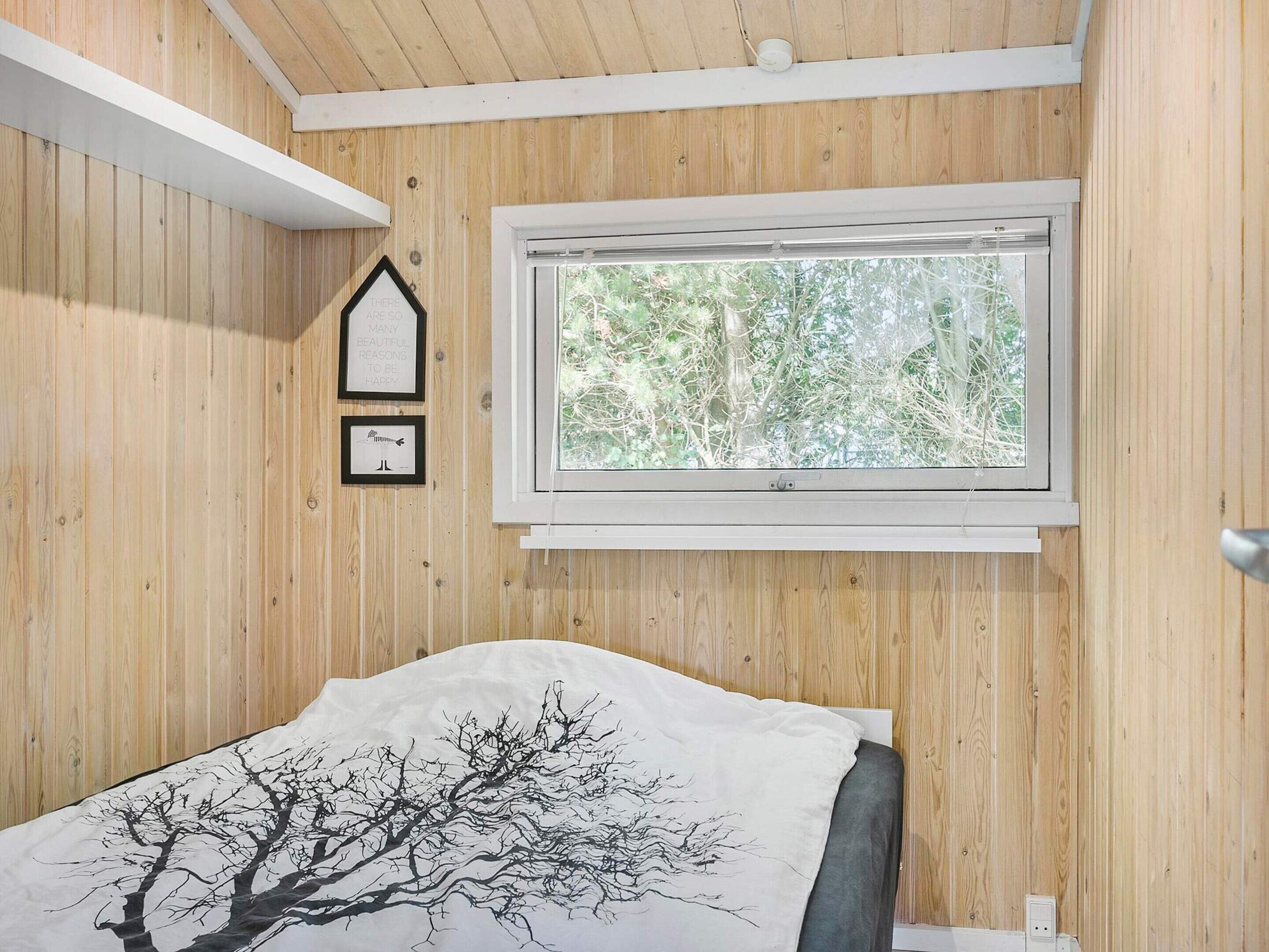 A bedroom with wooden walls, a window with a view of trees, a bed with a tree design on the pillow, and two framed pictures on the wall.