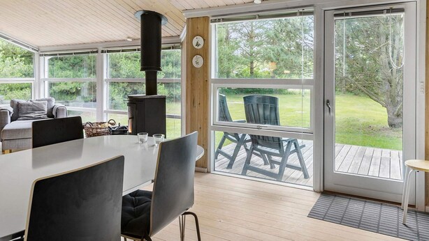 A modern interior with a large table and chairs, a wood-burning stove, and a sliding glass door leading to an outdoor deck. - 5 Star Holiday Home in Saltum (Saltum)
