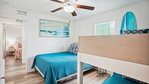 A bedroom with a bunk bed, a ceiling fan, a painting, and a surfboard on the wall. - Modern Coastal Condo - 4 Minute Walk to the Beach! (Madeira Beach)