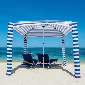 A beach umbrella with striped fabric and metal poles, two blue lounge chairs, and a clear view of the ocean and boats in the background.