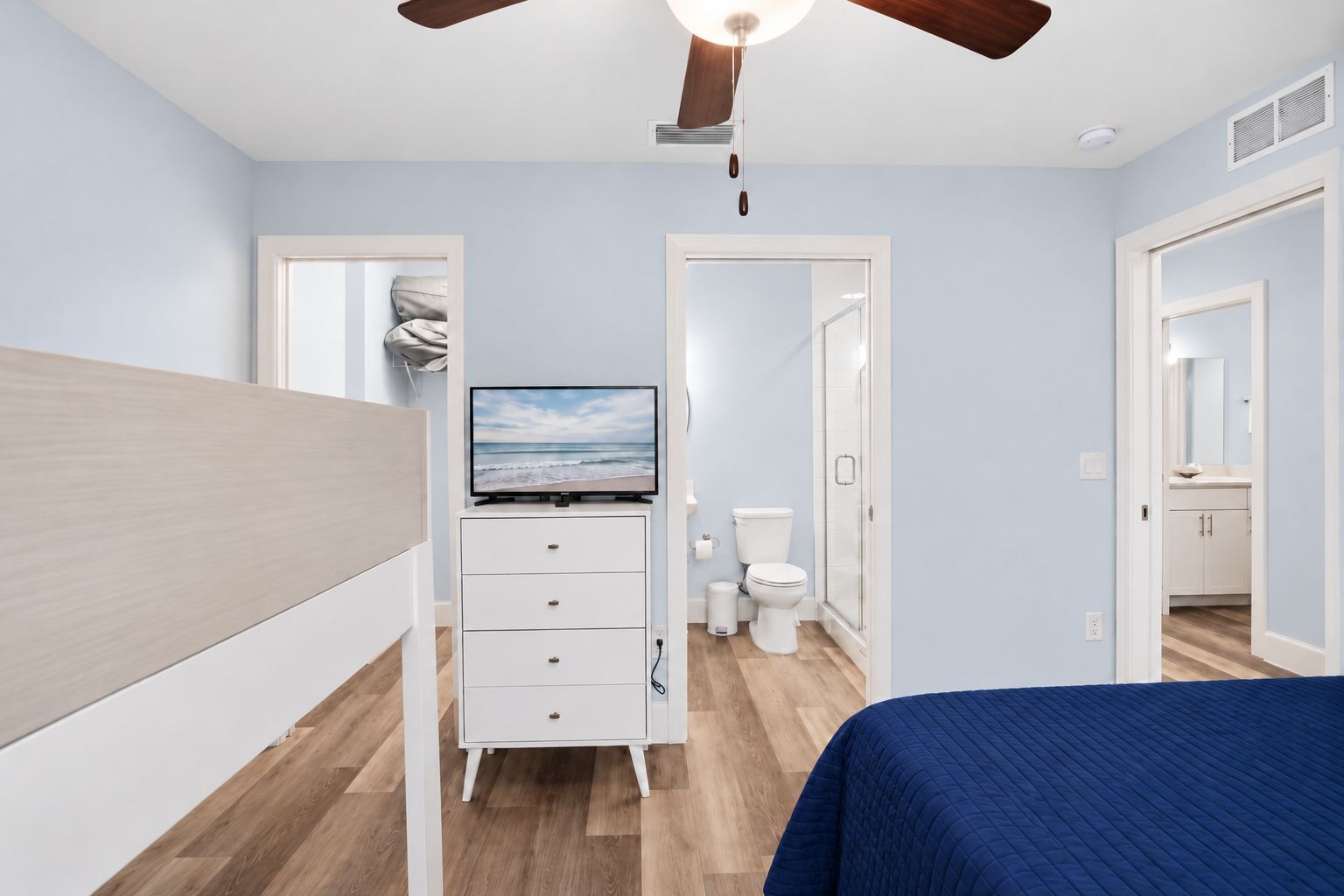 Un dormitorio con cama, televisor sobre un mueble, ventilador de techo y vista al baño.