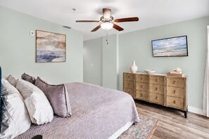 Une chambre à coucher comprenant un lit, une commode, un ventilateur de plafond et deux tableaux accrochés au mur.