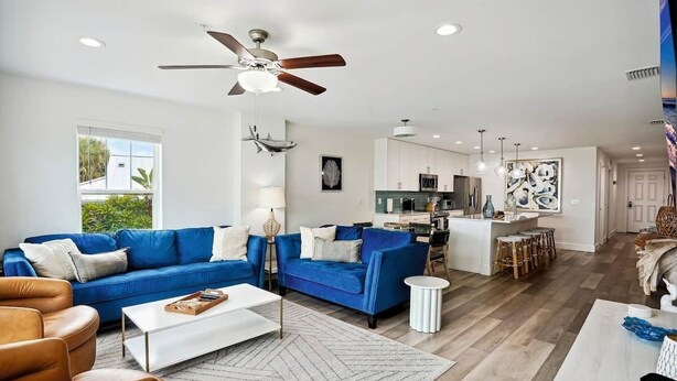 A spacious living room with a blue sofa, a coffee table, and a ceiling fan. - Modern Coastal Condo - 4 Minute Walk to the Beach! (Madeira Beach)
