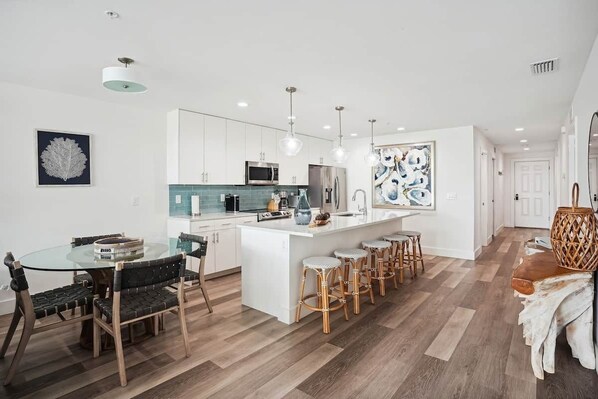 A modern kitchen with white cabinets, a central island with stools, and pendant lighting. - Modern Coastal Condo - 4 Minute Walk to the Beach! (Madeira Beach)