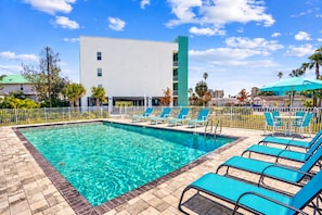 Une piscine rectangulaire à l’eau claire, entourée d’une terrasse carrelée. On y trouve plusieurs chaises longues bleues et une petite table avec un parasol bleu.