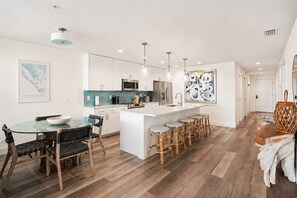 Una cocina moderna con armarios blancos, una isla central con taburetes y un comedor con mesa y sillas de vidrio.