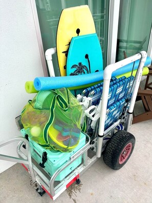 A beach cart with surfboards, a green mesh bag, and a blue chair.