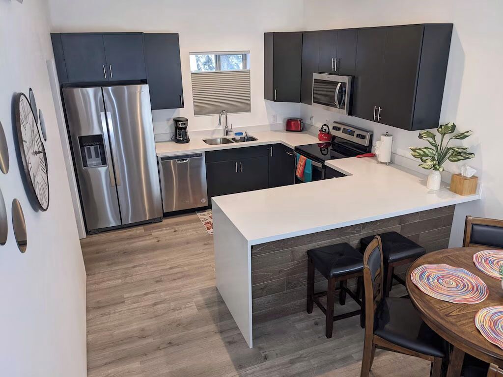 Een moderne keuken met donkere kastjes, roestvrijstalen apparatuur, een wit aanrecht en een houten eettafel met stoelen.