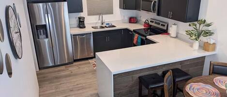 Een moderne keuken met donkere kastjes, roestvrijstalen apparatuur, een wit aanrecht en een houten eettafel met stoelen.