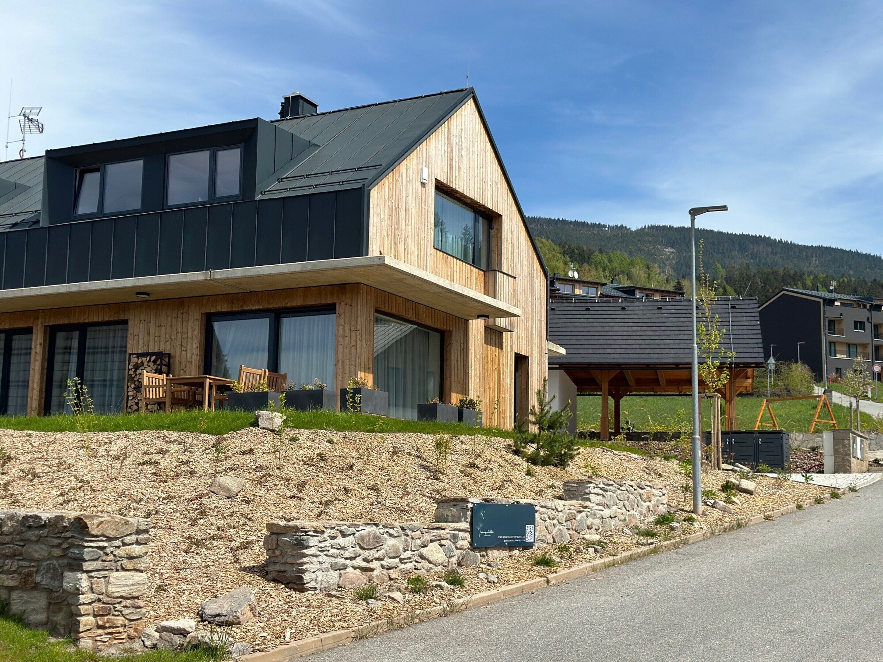 A modern two-story house with a wooden facade, large windows, and a stone retaining wall.