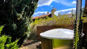 A hot tub in a garden with a wooden deck, surrounded by greenery and a clear sky.