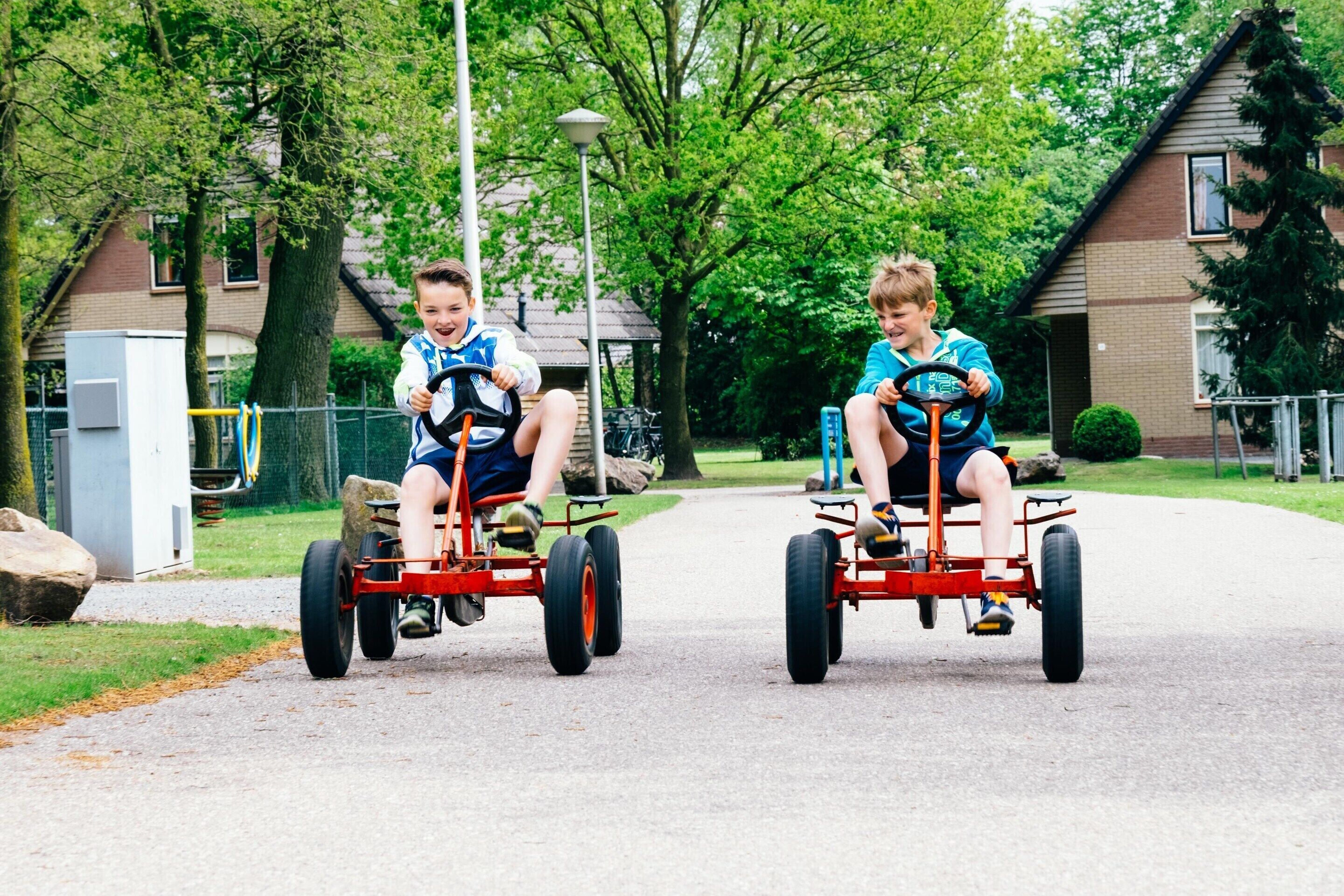 To børn kører gokarts på en belagt sti i et boligområde med huse og træer i baggrunden.
