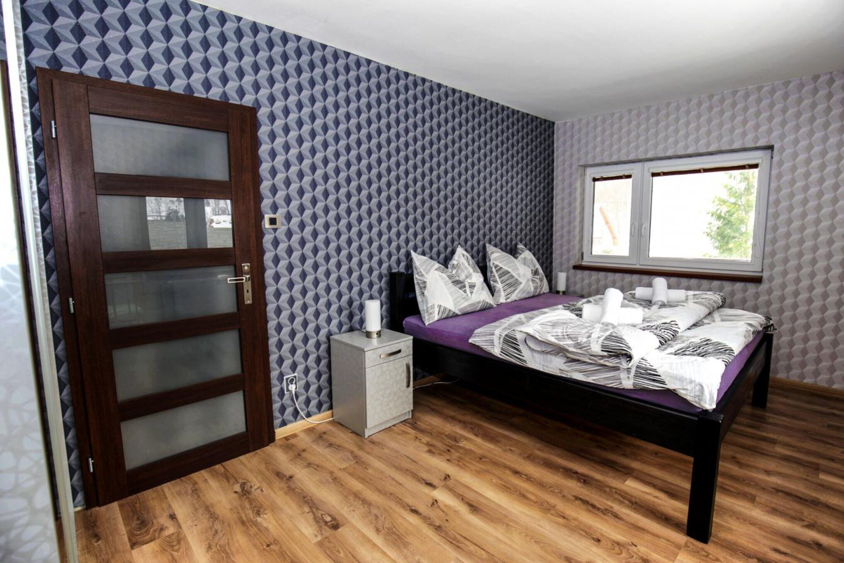 A bedroom with a wooden floor, a bed with a purple bedspread, a nightstand, a window, and patterned wallpaper.