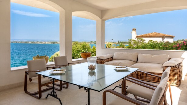 Ein sonnendurchflutetes Zimmer mit einem Glastisch, Stühlen und einer Couch. Es gibt große Fenster mit Blick auf das Meer und ein Gebäude gegenüber dem Wasser.