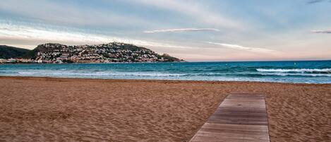 A sandy beach with a wooden walkway leading to the sea, overlooking a hillside with buildings.