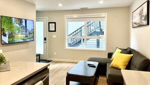 A modern living room with a large window, a flat-screen TV, a sofa with a yellow pillow, and a small table.
