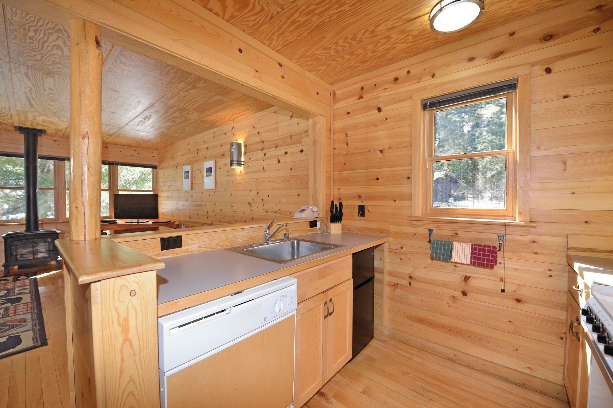 A wooden cabin interior with a kitchen area, including a sink, stove, and a window with a view of trees.