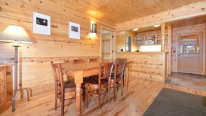A wooden interior with a dining table and chairs, a lamp, framed pictures on the wall, and a kitchen area with a sink and cabinets.