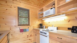 A compact kitchen with wooden walls, a stove, a microwave, and a window with blinds.