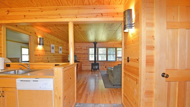 A cabin interior with wooden walls, a kitchen area with a sink and dishwasher, a fireplace, and a window with blinds. - Rustic Two Bedroom Cabin (Mazama)