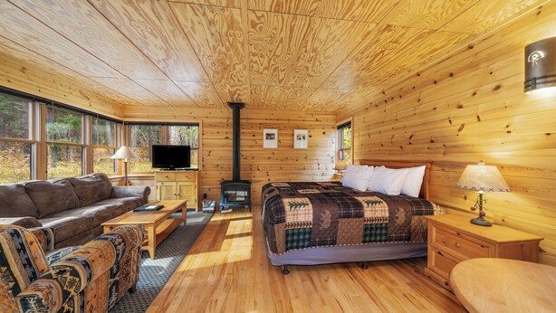 A cozy cabin interior with a wooden floor, a bed with a quilt, a sofa, a television, and a fireplace.