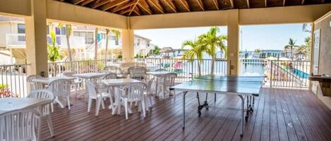 Une terrasse extérieure avec des tables et des chaises, une table de ping-pong, et une vue sur l’océan et les bâtiments en arrière-plan.