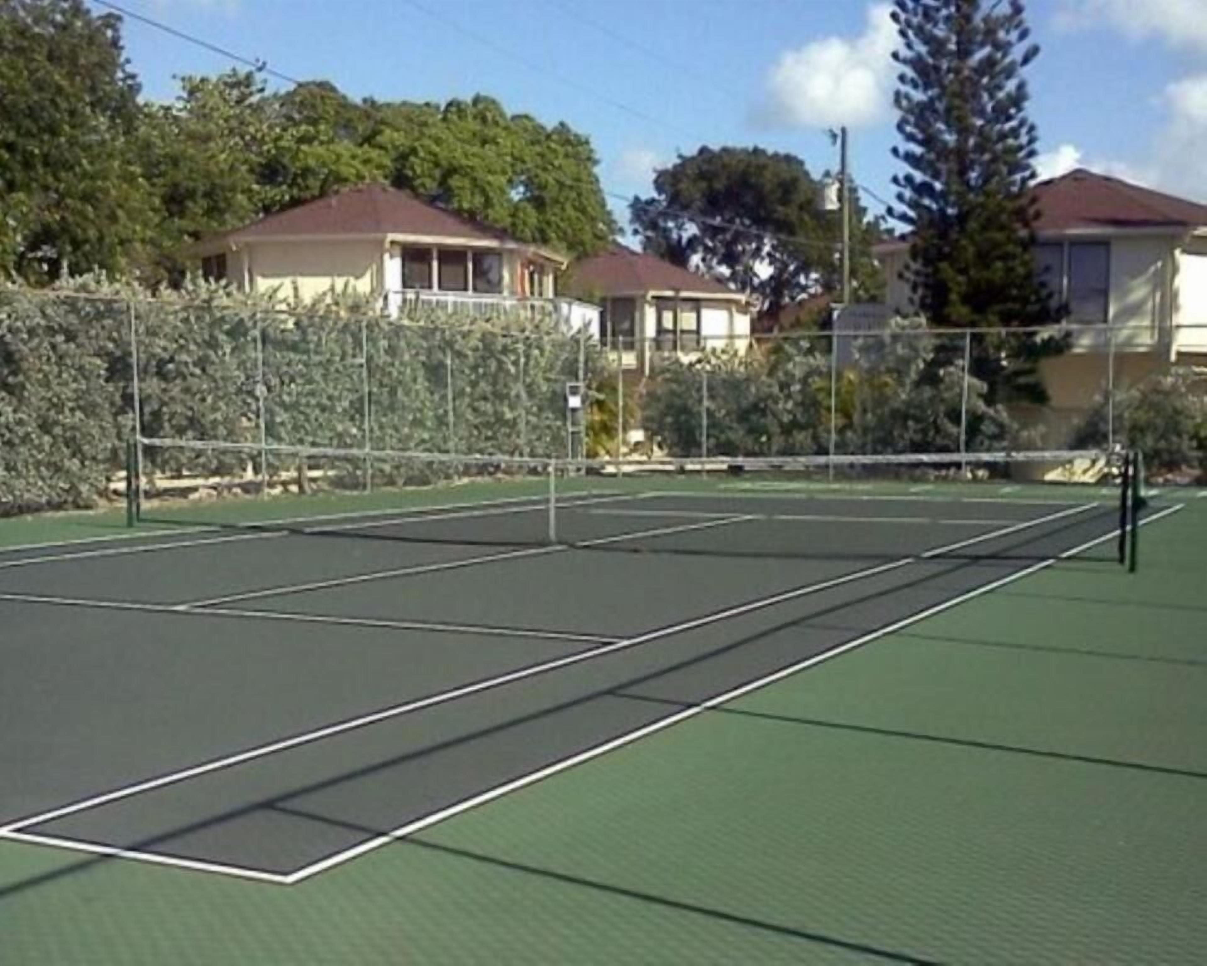 An outdoor tennis court with a net, surrounded by a fence and trees.