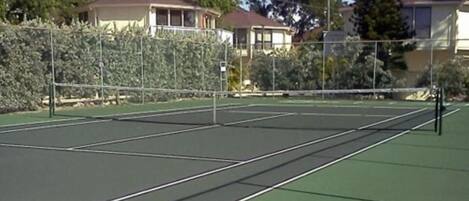 An outdoor tennis court with a net, surrounded by a fence and trees.