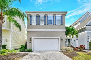 A two-story house with a white garage door, a balcony with blue shutters, and a well-maintained garden with various plants and a palm tree.