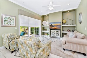 A spacious living room with a sectional sofa, a chaise lounge, and a flat-screen TV. There is a ceiling fan, a fireplace, and a large window with a view of the outdoors.